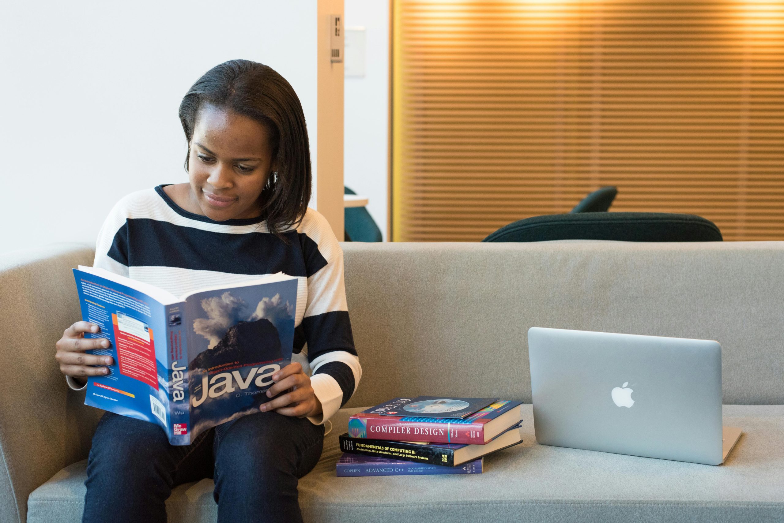A woman studying Java programming on a sofa with a MacBook and other computer books nearby.